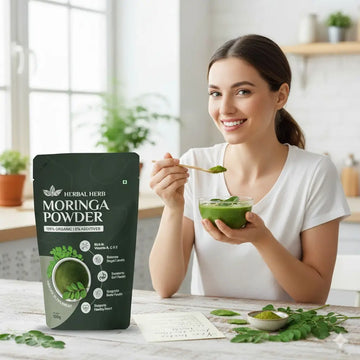 Woman holding a bowl of green smoothie next to a package of Herbal Herb Moringa Powder in a bright kitchen.